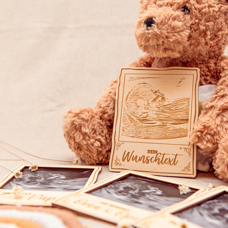 May include: A wooden photo frame with a brown teddy bear holding it. The frame has a cutout of an ultrasound image and the text "Dein Wunschtext".