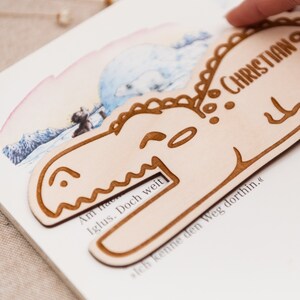 May include: A light brown wooden bookmark shaped like a dinosaur with the text "CHRISTIAN" on the top and "Ich kenne den Weg dorthin." on the bottom.