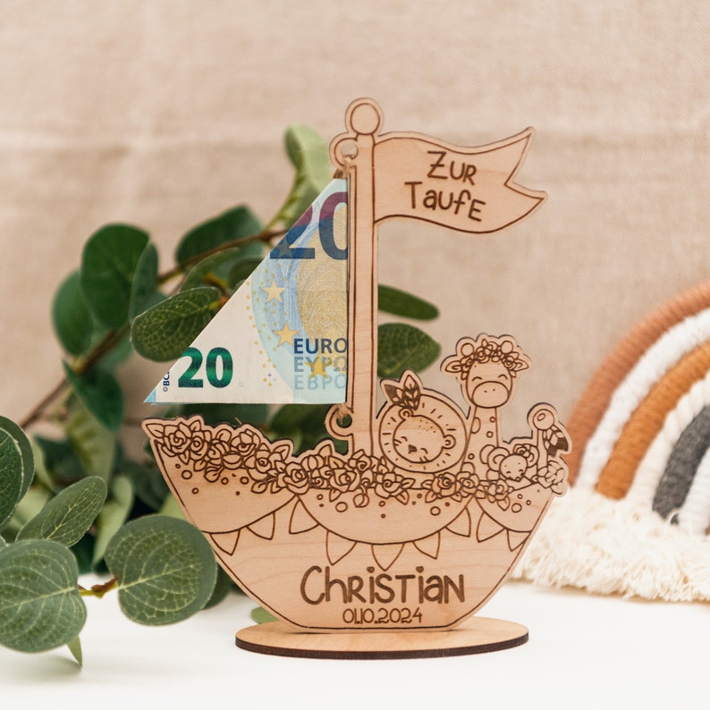 Puede incluir: Hucha de madera en forma de velero con una jirafa, un le&oacute;n y un rat&oacute;n. La vela est&aacute; hecha de un billete de 20 euros. La caja est&aacute; grabada con el texto "Zur Taufe" y "Christian 01.01.2024".