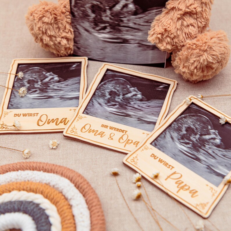 May include: Three wooden photo frames with ultrasound images and personalised text. The frames are decorated with floral designs and the text reads "Du wirst Oma", "Ihr werdet Oma & Opa", and "Du wirst Papa".