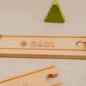 May include: A light brown wooden train track piece with the name "KARL" engraved in a serif font, and a small illustration. A green, triangular wooden tree is in the background. The track is made of wood.