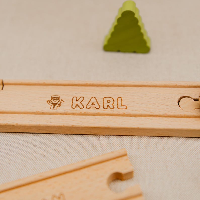 May include: A light brown wooden train track piece with the name "KARL" engraved in a serif font, and a small illustration. A green, triangular wooden tree is in the background. The track is made of wood.