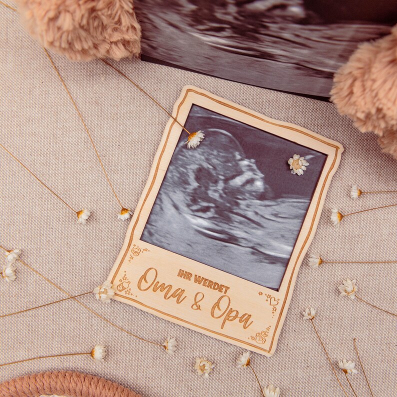 May include: A wooden frame with a black and white ultrasound image inside. The frame is decorated with small white flowers and the text "Ihr werdet Oma & Opa".