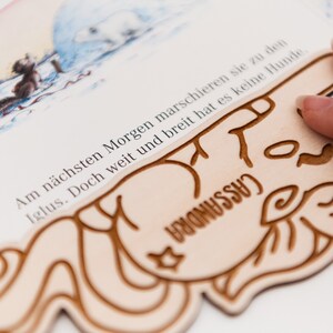 May include: A close-up of a wooden book with a carved illustration of a dog. The book is open to a page with text in German that reads: "Am n&auml;chsten Morgen marschieren sie zu den Iglus. Doch weit und breit hat es keine Hunde." The text is partially obscured by the dog illustration.