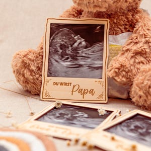 May include: Wooden photo frame with a black and white ultrasound image inside. The frame has a light brown wood finish and says "Du wirst Papa" in a decorative font.