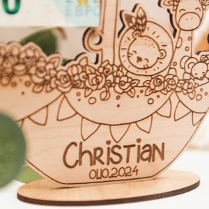 Puede incluir: Corte de madera de un barco con un le&oacute;n, una jirafa y un mono a bordo. El barco est&aacute; decorado con flores y el nombre "Christian" y la fecha "01.10.2024" est&aacute;n grabados en la parte inferior.