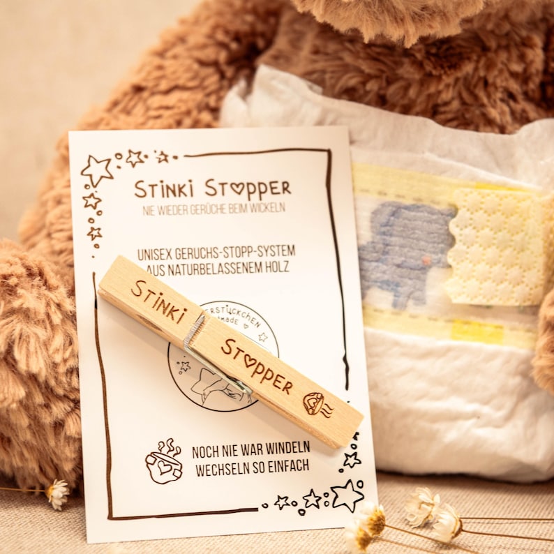 May include: A wooden clothespin with the words "Stinki Stopper" printed on it. The clothespin is sitting on a white card with the text "Stinki Stopper, Ne wieder Ger&uuml;che beim Wickeln, Unisex Geruchs-Stopp-System aus Naturbelassenem Holz, Noch nie war Windeln wechseln so einfach".