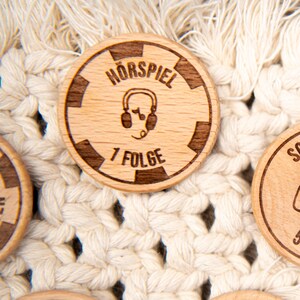 May include: A wooden token with a black and white design. The token has the text "H&ouml;rspiel 7 Folge" and a graphic of headphones with a musical note.