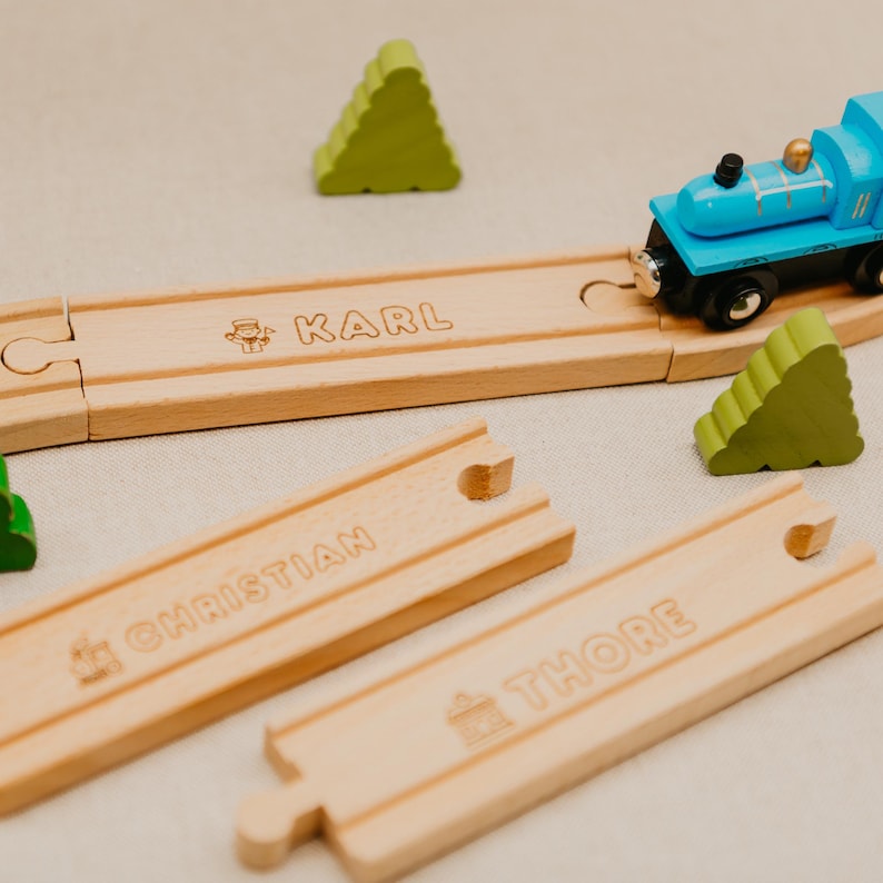 May include: A wooden toy train set featuring a blue train, wooden tracks, and green tree-shaped blocks. The tracks are engraved with the names "KARL", "CHRISTIAN", and "THORE". The train is on the track, ready to play.