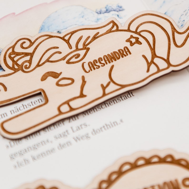 May include: A wooden unicorn bookmark with the name "CASSANDRA" engraved on it. The bookmark is light brown and has a star on the unicorn's horn.