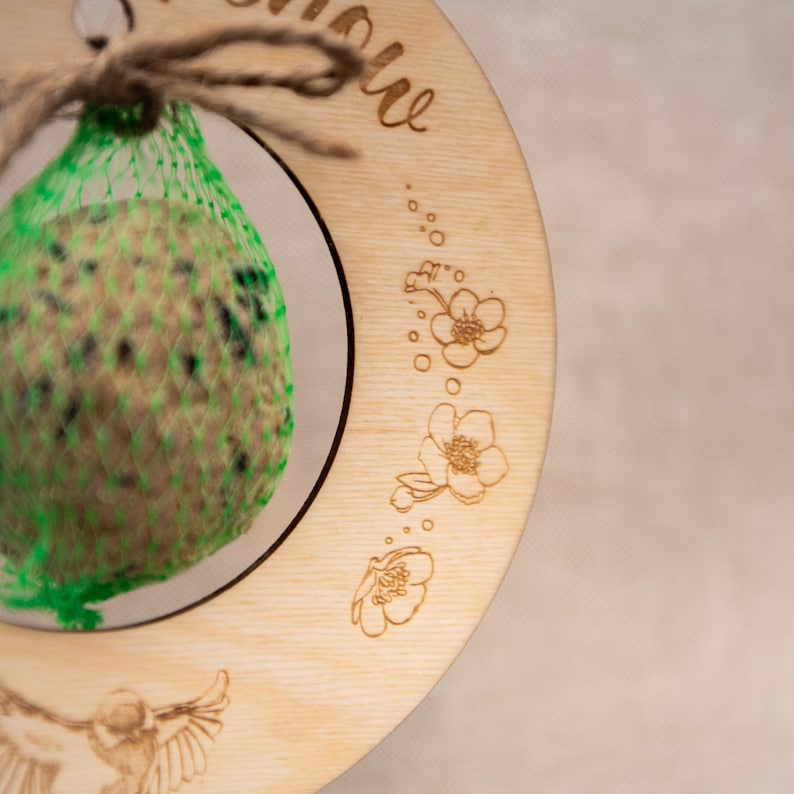 Pode incluir: Um comedouro de p&aacute;ssaros de madeira com um saco de malha verde cheio de sementes para p&aacute;ssaros. O comedouro &eacute; decorado com um desenho floral e as palavras "Bem-vindos, passarinhos".