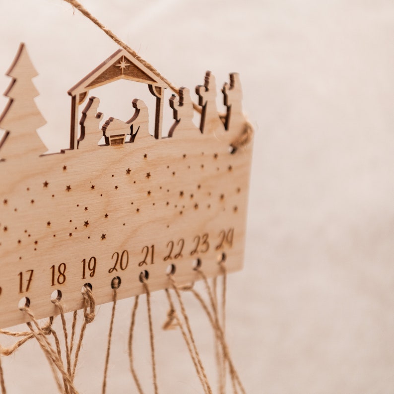 May include: A wooden advent calendar with a nativity scene design. The calendar has 24 numbered slots with twine strings hanging from each slot. The numbers 17 through 24 are visible.