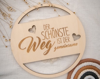 May include: A round wooden sign with the text "Der schönste Weg ist der gemeinsame" (The most beautiful path is the shared one) in German. The sign has two cut-out hearts and a fringe detail.