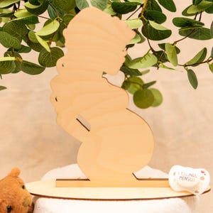 May include: A wooden silhouette of a pregnant person, standing on a round wooden base. The figure is light brown and has a simple, elegant design. A small pacifier with text is visible on the base. Green leaves are in the background.