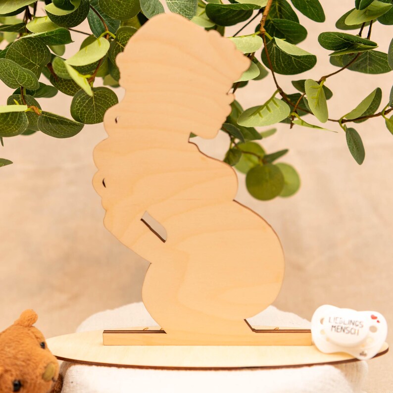May include: A wooden silhouette of a pregnant person, standing on a round wooden base. The figure is light brown and has a simple, elegant design. A small pacifier with text is visible on the base. Green leaves are in the background.