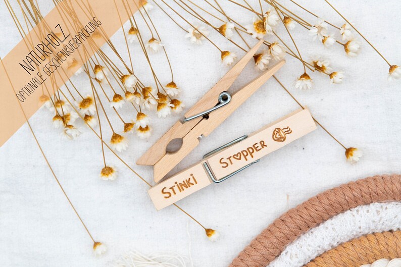 May include: Two wooden clothespins with the words "Stinki" and "Stopper" engraved on them. The clothespins are surrounded by dried white flowers and a brown and white woven rug.