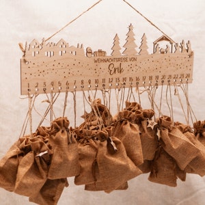 May include: Wooden Christmas advent calendar with a winter village scene and the text "WEIHNACHTSREISE VON Erik".  The calendar has 24 small burlap bags hanging from it, each with a number.