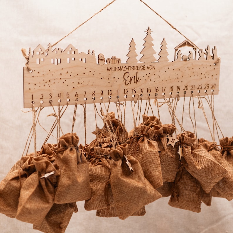 May include: Wooden Christmas advent calendar with a winter village scene and the text "WEIHNACHTSREISE VON Erik".  The calendar has 24 small burlap bags hanging from it, each with a number.