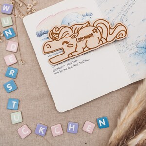 May include: A wooden unicorn bookmark with the name "Cassandra" engraved on it. The bookmark is lying on a page of a book with German text.