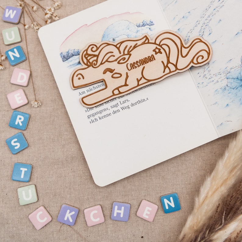 May include: A wooden unicorn bookmark with the name "Cassandra" engraved on it. The bookmark is lying on a page of a book with German text.
