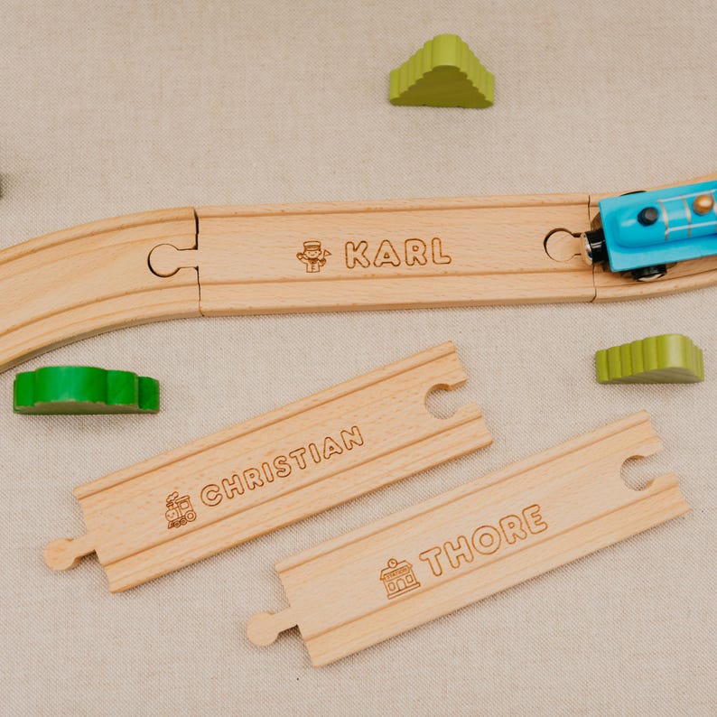 May include: Wooden toy train track pieces engraved with the names "Karl", "Christian", and "Thore". A blue toy train is on the track. Green, tree-shaped wooden blocks are scattered around the track.