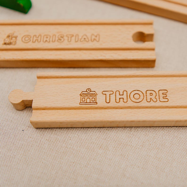 May include: Wooden train track pieces with engraved names "CHRISTIAN" and "THORE". The "THORE" piece includes a station icon. The tracks are light brown and feature grooves for train wheels. A fun toy for children.