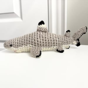 May include: A crocheted shark toy in shades of gray, white, and black. The shark has a detailed, textured body, fins, and tail. The toy is designed to resemble a shark, with a small mouth and eyes. The shark is approximately 10 inches long.