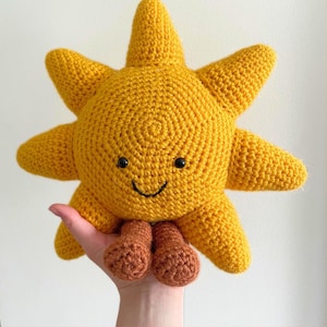 Op de afbeelding: Een handgemaakt, gehaakt zonnespeelgoed in helder geel met een lachend gezicht en bruine voetjes. De zon heeft acht punten en wordt in een hand gehouden. Het speelgoed is zacht en lijkt van garen te zijn gemaakt.