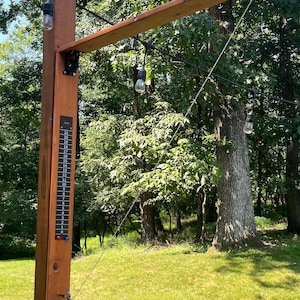 May include: A wooden outdoor shower fixture with a brown post and a horizontal wooden arm. The fixture has a temperature gauge, a shower head, and string lights. The background features green grass and trees.
