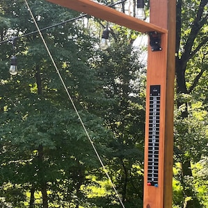 May include: A wooden outdoor game set with a brown post, a wooden arm, and hanging light bulbs. A scoring chart is attached to the post. A rope and ring are visible, ready for play. The background is a green lawn and trees.