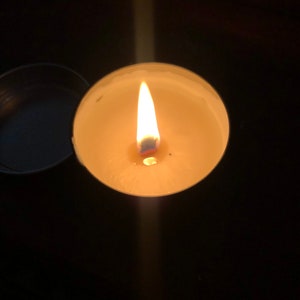May include: A lit, round candle in a white container. The flame is bright yellow and orange, with a small pool of melted wax visible. The candle sits on a dark surface, with a partially visible, open, round container to the left.
