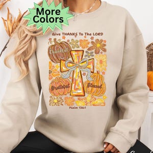 May include: Sand-colored sweatshirt with a graphic featuring a cross, pumpkins, and floral elements. Text includes "Give Thanks To The Lord," "Thankful," "Grateful," and "Blessed." The design has a vintage, autumnal aesthetic. The words "More Colors" are also visible.