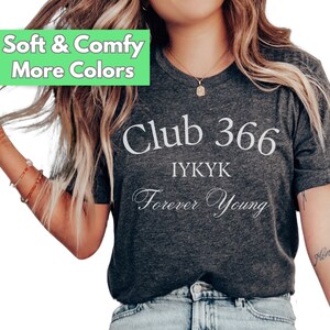 May include: A dark grey heather t-shirt with the text "Club 366 IYKYK Forever Young" printed in white. The model is wearing blue jeans.