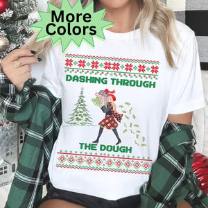 May include: White t-shirt with a festive design. The shirt features the text "Dashing Through The Dough" in green, with a graphic of a woman carrying gifts and money. The design includes red and green snowflake patterns, and a Christmas tree.