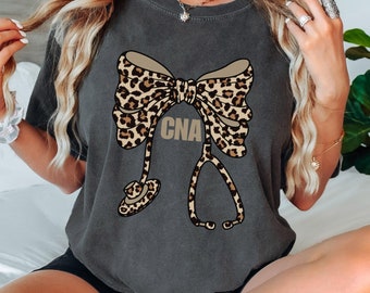 May include: Dark gray t-shirt featuring a leopard print bow and stethoscope design. The bow has a tan background with brown spots. The word "CNA" is printed in tan. The shirt is a comfortable, casual style.