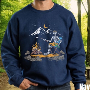May include: A navy blue sweatshirt with a graphic of a skeleton roasting a marshmallow over a campfire. The skeleton is sitting in a camping chair and holding a beer. The graphic is set against a backdrop of a mountain range and a crescent moon.