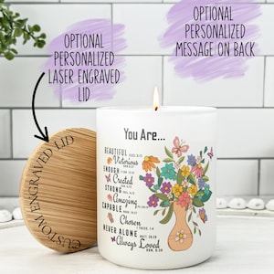 May include: A white candle with a floral design and the words "You Are..." printed on it. The candle has a wooden lid with the words "Custom Engraved Lid" on it. The candle is lit and has a soft glow.
