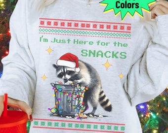 May include: A light gray sweatshirt with a festive design. It features a raccoon wearing a Santa hat, standing by a trash can with Christmas lights. The text reads, "I'm Just Here for the SNACKS." Decorative borders and star accents complete the design.