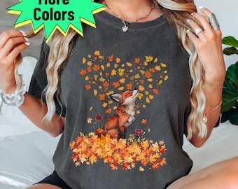 May include: Dark grey t-shirt featuring a fox illustration surrounded by autumn leaves. The fox is depicted with orange and brown fur, looking upwards. The leaves form a heart shape above the fox. The words "More Colors" are in a green starburst.