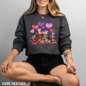 May include: Dark grey sweatshirt with a Valentine's Day theme. The design shows dachshund dogs, heart-shaped balloons, and the word "LOVE". The phrase "Free Kisses" is also visible. A comfortable, casual style of clothing.