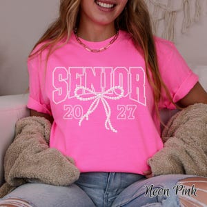 Puede incluir: Una camiseta de manga corta color rosa ne&oacute;n con la palabra SENIOR en letras blancas de bloque, con los n&uacute;meros 20 y 27 a los lados. Un gr&aacute;fico blanco de un lazo hecho de perlas est&aacute; centrado debajo de la palabra SENIOR, sobre los n&uacute;meros.