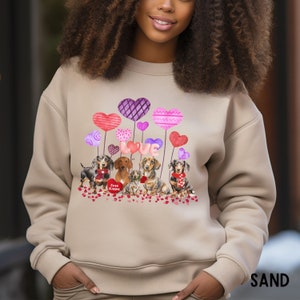May include: Sand-coloured sweatshirt with a Valentine's Day design. The graphic features dachshunds, heart-shaped balloons, and the word "LOVE." The design also includes the text "Free Kisses" on a heart.