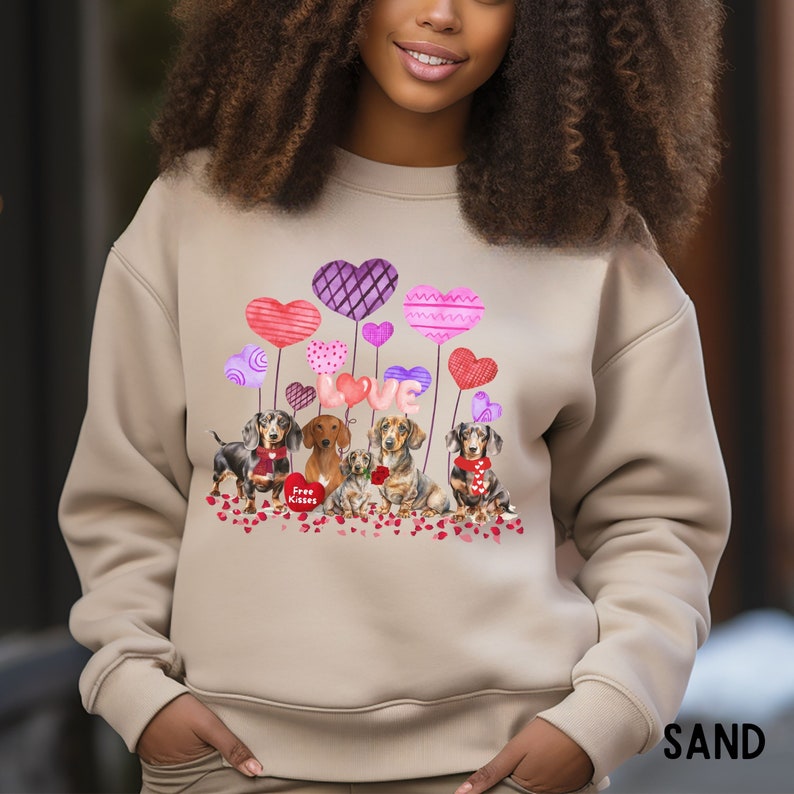 May include: Sand-coloured sweatshirt with a Valentine's Day design. The graphic features dachshunds, heart-shaped balloons, and the word "LOVE." The design also includes the text "Free Kisses" on a heart.