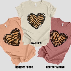 May include: Three t-shirts with a tiger stripe heart design. The shirts are in the colors natural, heather peach, and heather mauve.