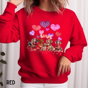 May include: Red sweatshirt with a graphic of dachshunds, heart-shaped balloons, and the word "LOVE". The design includes the text "Free Kisses". A colourful and festive design for dog lovers.