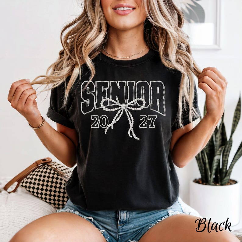 Puede incluir: Una camiseta negra de manga corta con la palabra SENIOR en letras blancas de bloque y un gr&aacute;fico de lazo estilo perla blanca centrado debajo del texto, flanqueado por los n&uacute;meros 20 y 27 en blanco en la parte frontal de la prenda.