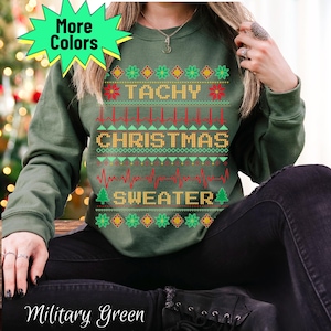 May include: Military green sweatshirt with the text "TACHY CHRISTMAS SWEATER" in a festive design. The design includes red and green elements, including a heartbeat line, and Christmas tree graphics. The words "More Colors" are in a starburst.