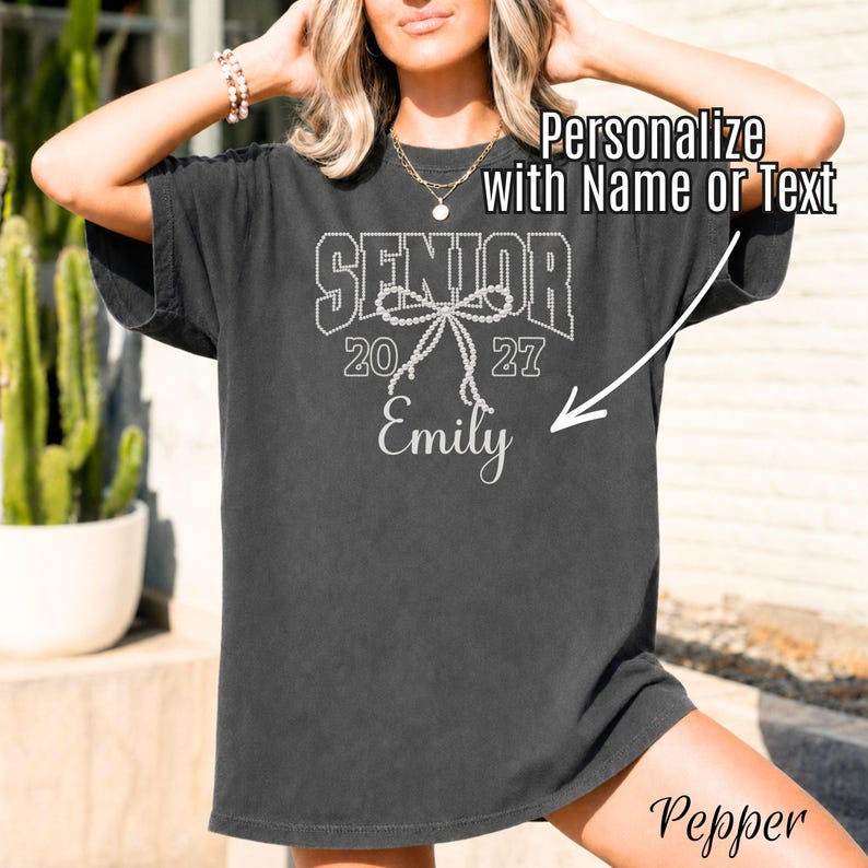 Puede incluir: Una camiseta gris carb&oacute;n de manga corta con un gr&aacute;fico blanco que incluye el texto SENIOR 2027, una ilustraci&oacute;n de un lazo estilo perlas y el nombre Emily. El dise&ntilde;o incluye una flecha que apunta al nombre con el texto Personalize with Name or Text.
