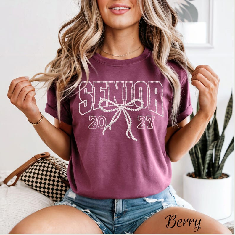 Puede incluir: Una camiseta de manga corta color baya con el texto SENIOR 2027 en una fuente blanca de puntos. Un gr&aacute;fico grande de un lazo blanco de puntos est&aacute; centrado entre los n&uacute;meros. La camiseta tiene cuello redondo y un corte holgado de algod&oacute;n.