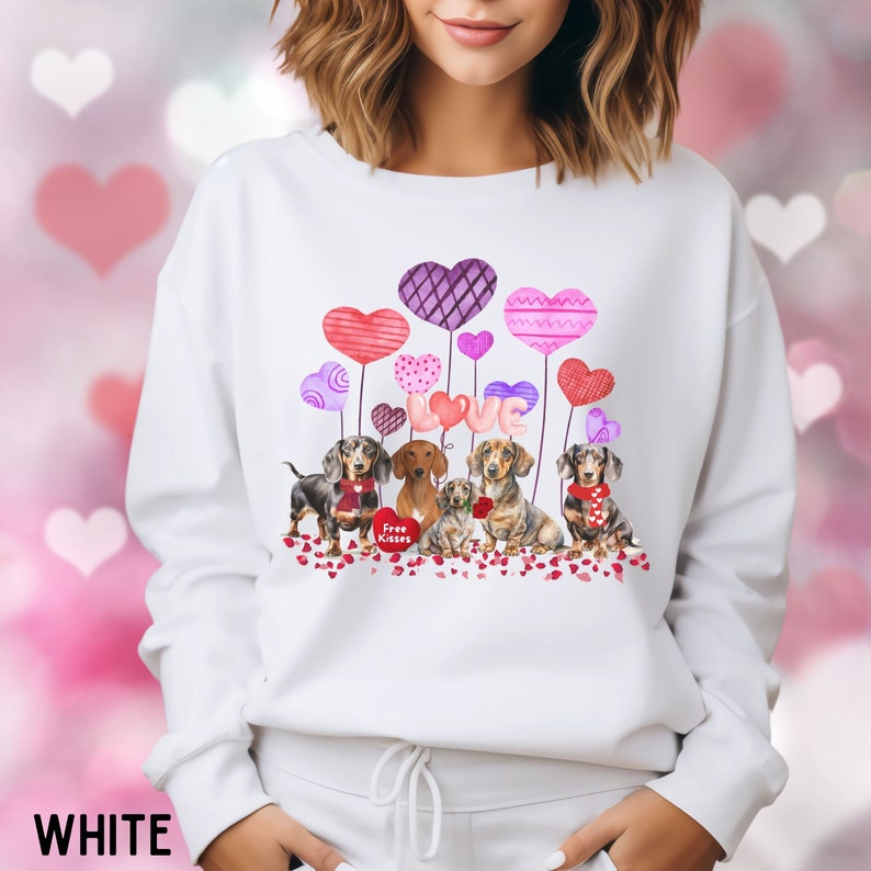 May include: White sweatshirt with a Valentine's Day theme, showcasing dachshunds, heart-shaped balloons, and the word "LOVE". The design incorporates red, pink, and purple hearts, and the text "Free Kisses".
